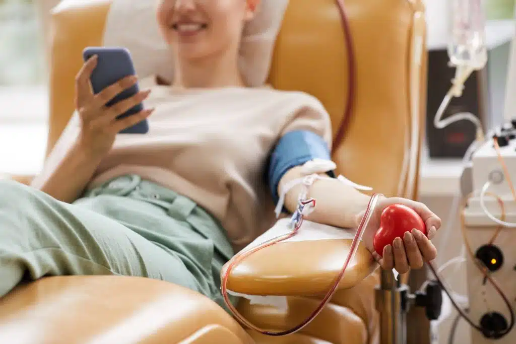 young woman sitting in a chair looking at her phone while giving blood
