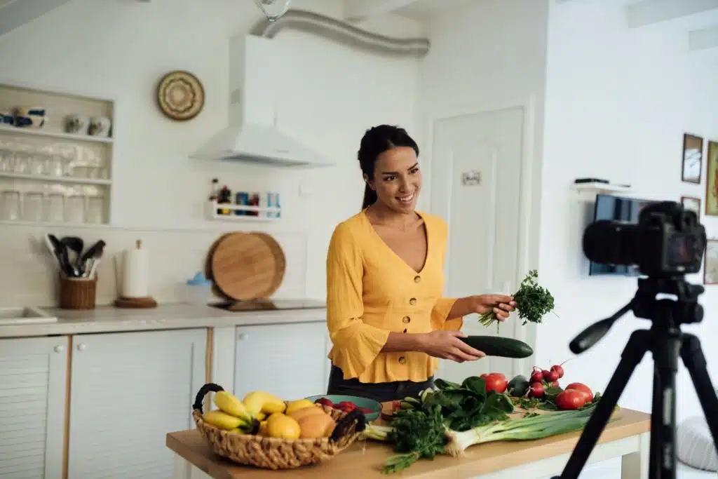 woman standing in a kitchen filming a video while holding vegetables in her hands