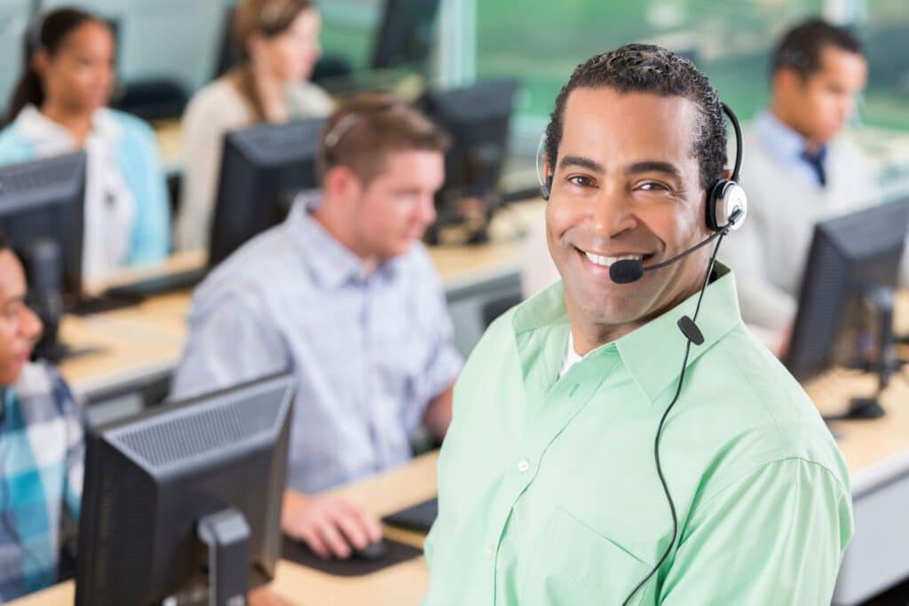 Man with a headset on smiling for the camera with call center employees in the background