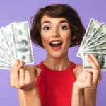 Happy Pretty brunette woman showing money while looking at the camera over purple background - making six figures