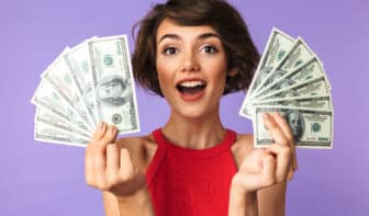 Happy Pretty brunette woman showing money while looking at the camera over purple background - making six figures
