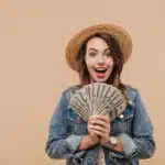 Portrait of a smiling young girl in summer clothes showing money banknotes and looking at camera isolated over beige background for the concept "What is passive income?"