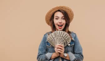 Portrait of a smiling young girl in summer clothes showing money banknotes and looking at camera isolated over beige background for the concept "What is passive income?"
